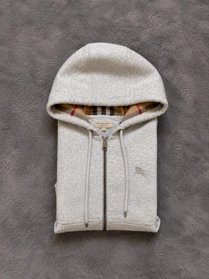 Grå zip hoodie från Burberry - Snygg grå hoodie från Burberry med dragkedja framtill och klassiskt rutigt foder i huvan. Tillverkad i mjuk bomullsblandning, har två fickor fram och diskret Burberry-logga broderad på bröstet. Perfekt för en avslappnad men stilren look.