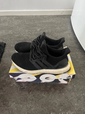 Svarta Adidas Ultra Boost sneakers - Säljer ett par svarta Adidas Ultra Boost sneakers med stickad ovandel och vita Boost-sulor. Skorna har svarta plastdetaljer på sidorna och hälkappa samt klassiska tre ränder. Perfekta för dig som gillar stilrena och bekväma sneakers.
