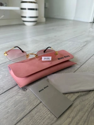 Miu Miu rosa solglasögon MU A54S ZVN70O - Säljer ett par trendiga solglasögon från Miu Miu med tunna guldfärgade bågar och rosa glas. Glasögonen har en elegant, kantig form och kommer med ett matchande rosa fodral och putsduk. Perfekt accessoar för dig som vill sticka ut i sommar .Miu Miu MU A54S ZVN70O