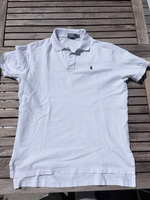 Vit pikétröja från Ralph Lauren - Klassisk vit pikétröja från Ralph Lauren med korta ärmar och broderad logga på bröstet. Tröjan har krage och knäppning framtill. Perfekt för en clean och stilren look.