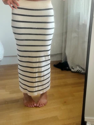 Randig långkjol med slits - Säljer en snygg långkjol i vitt med svarta horisontella ränder. Kjolen har en hög midja och en slits på sidan som ger en trendig touch. Materialet känns mjukt och stretchigt, perfekt för en chill och stilren look.