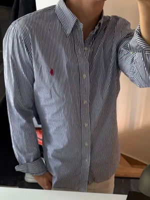 Randig skjorta från Polo Ralph Lauren - Klassisk blåvit randig skjorta från Polo Ralph Lauren med röd broderad logga på bröstet. Skjortan har lång ärm, knappar framtill och button-down krage. Tillverkad i bomull för en skön och stilren look.