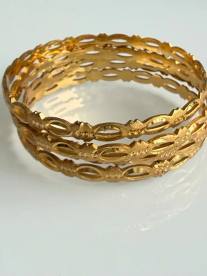 Guldfärgade utskurna armband - Säljer ett set med tre guldfärgade armband i metall med utskurna detaljer och ovala former. Armbanden har en tunn och lätt design med dekorativa mönster som ger en trendig och elegant look. Perfekta att bära tillsammans eller var för sig för en snygg stil. Två små skråmor syns på bild 3. Inget som syns på långt håll. 