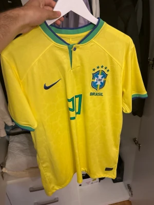 Brasilien matchtröja Nike Vini Jr - Gul Brasilien matchtröja från Nike med gröna och blå detaljer. Nummer 20 och VINI JR på ryggen, CBF-emblem och Nike-logga på bröstet. Tillverkad i lätt och ventilerande material, perfekt för fotboll eller träning.