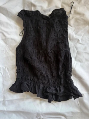 Svart ärmlös blus med volang - Svart ärmlös blus med smockad struktur och volangkant nedtill. Toppen har en lätt och luftig känsla, perfekt för varma dagar. Snygg att styla med både jeans och kjol.