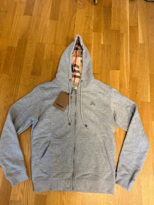 Grå zip hoodie från Burberry - Snygg grå hoodie från Burberry med dragkedja och klassiskt rutigt foder i huvan. Hoodien har två fickor fram, justerbar dragsko i huvan och diskret Burberry-logga på bröstet. Tillverkad i mjuk bomullsblandning för extra komfort.