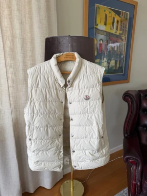 Vit dunväst från Moncler - Snygg vit dunväst från Moncler med ståkrage och tryckknappar framtill. Västen har Moncler-logga på bröstet och är tillverkad i ett glansigt material med certifierat dunfoder. Perfekt för lager-på-lager och ger en clean look.