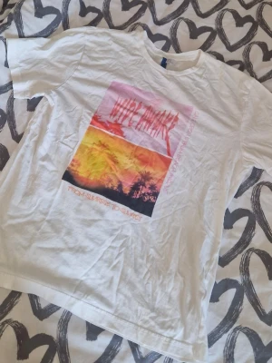 Vit t-shirt med tryck Wide Awake - Vit t-shirt med färgglatt tryck framtill som visar palmer och solnedgång, samt texten 'Wide Awake' och 'From Sunrise to Sunset'. Skön passform och klassisk rund hals. Perfekt för en chill och avslappnad stil.