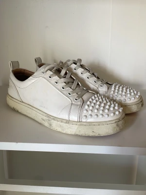 Vita Louboutins  - Unika vita sneakers från Christian Louboutin med coola nitar på tån och klassisk låg siluett. Skorna är gjorda i läder och har vita snören samt en rejäl sula. Perfekta för dig som vill sticka ut med en edgy och exklusiv look. Dem ser rätt slitna ut på bilderna men jag kan tvätta rent dem innan dem skickas. Dem kommer med box 