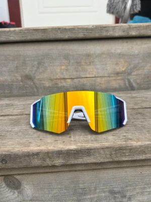 Vita sportiga solglasögon med spegelglas - Säljer ett par riktigt snygga sportiga solglasögon med vit båge och stora spegelglas i gult, blått och grönt. Glasen har en futuristisk, kantig form och ger en cool vibe till din outfit. Perfekta för dig som vill sticka ut i sommar!