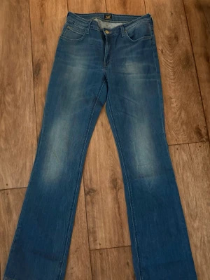Blå bootcut jeans från Lee - Säljer ett par klassiska blå bootcut jeans från Lee med fem fickor och snygga slitningar framtill. Jeansen har normal midja och Lee-lapp bak i linningen. Perfekta för dig som gillar en avslappnad men ändå trendig look. W29 L34