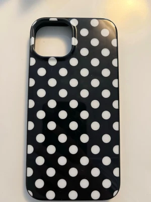 Prickigt mobilskal - Snyggt svart mobilskal för iPhone 13 och 14 med vita polkaprickar. Skalets yta är blank och det har en öppning för kameran. Passar perfekt för dig som vill ha ett trendigt skal till din mobil. Materialet känns tåligt och skalet är rätt så bra kvalitet men har några märken på ovansidan men syns inte mycket. Mobilskalet har MagSafe  (magnetisk anslutning)