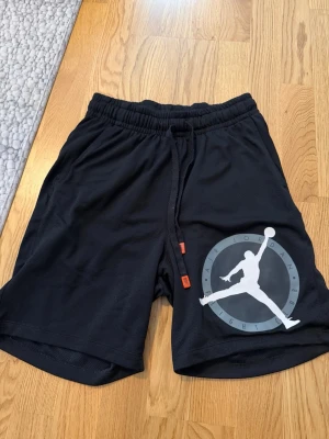 Svarta Jordan shorts med logga - Säljer ett par svarta Jordan shorts med stor vit Jumpman-logga på ena benet. Shortsen har elastisk midja med snörning och är gjorda i ett lätt, ventilerande syntetmaterial. Perfekta för sport eller chill.