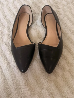 Svarta spetsiga ballerinaskor - Snygga svarta ballerinaskor med spetsig tå och öppen sida. Skorna har en stilren design och är tillverkade i slätt material med beige innersula. Lite lätt slitna (se sista bilden)