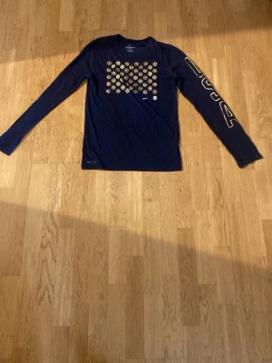 Blå långärmad Nike USA t-shirt - Mörkblå långärmad t-shirt från Nike med guldiga basketbollar på bröstet och stor 'USA' text på ena ärmen. Tillverkad i Dri-FIT material som andas, perfekt för sportiga dagar. USA-basketlogga och Nike swoosh på framsidan. Storleken är S och är knappast använd 