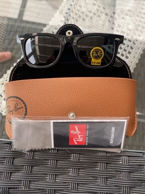 Svarta Ray-Ban Wayfarer solglasögon - Klassiska svarta Ray-Ban Wayfarer solglasögon med blank finish och ikonisk form. Kommer med original brunt fodral, putsduk och etikett. Perfekta för dig som vill ha en tidlös och stilren accessoar till sommaren.