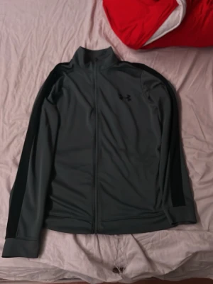 Svartgrå träningsjacka Under Armour - Snygg svartgrå träningsjacka från Under Armour med hel dragkedja och högt krage. Jackan har svarta detaljer längs ärmarna och är tillverkad i ett stretchigt funktionsmaterial som passar perfekt till träning eller uppvärmning.