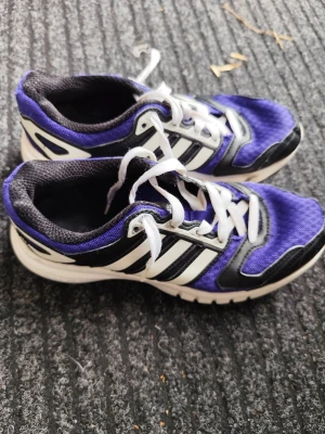 Lila Adidas sneakers med vita ränder - Snygga sneakers från Adidas i lila mesh med svarta och vita detaljer samt klassiska tre ränder på sidan. Skorna har vita snören och en vit sula. Perfekta för dig som gillar sportig stil och vill sticka ut lite extra.