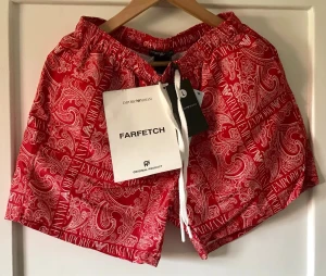 Röda badbyxor Emporio Armani paisley - Snygga röda badbyxor från Emporio Armani med paisleymönster och vita loggor längs benen. Resår i midjan och vita dragsnören framtill. Perfekta för stranden eller poolen, riktigt trendig vibe.