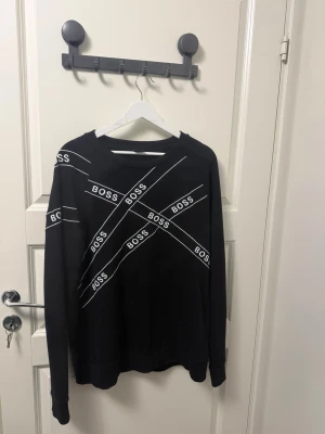 Svart sweatshirt från BOSS - Cool svart sweatshirt från BOSS med vita diagonala logor över hela framsidan. Tröjan har rund hals och långa ärmar, perfekt för en avslappnad och trendig look. Materialet ser ut att vara mjukt och bekvämt, idealiskt för chill dagar.