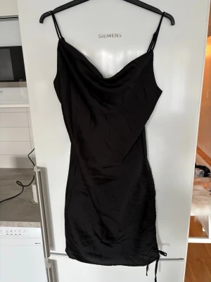 Svart satin klänning med snörning - Svart kort klänning i satin med tunna axelband och draperad urringning. Klänningen har snörningsdetalj på sidan som ger en snygg passform och trendig look. Perfekt för dig som gillar stilrena och eleganta plagg.