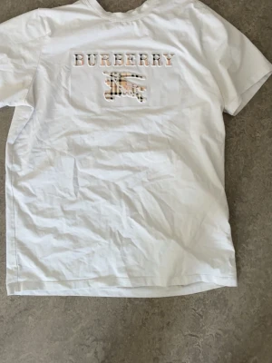 Vit  t-shirt med logga - Snygg vit t-shirt  med klassisk logga och riddare i beige, svart och röd rutig design på bröstet. T-shirten har rund hals och korta ärmar, perfekt för en clean och stilren look.