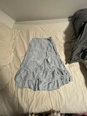 Blommig blå omlottkjol med volang - Söt blå omlottkjol med vitt blommönster och volangkant. Kjolen har knytband i midjan och är gjord i ett lätt och luftigt material, perfekt för varmare dagar. Passar dig som gillar romantisk och avslappnad stil.