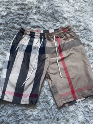 Beige-rutiga shorts med snörning - Säljer ett par snygga shorts med klassiskt beige, svart, vit och röd rutigt mönster. De har elastisk midja med vit snörning och sidofickor. Perfekta för sommaren och riktigt sköna att bära till vardags eller när du chillar.