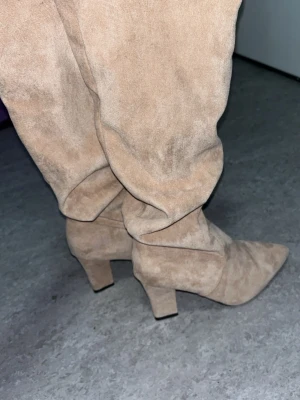 Beige mockastövlar med klack - Snygga beige stövlar i mocka-look med hög klack och spetsig tå. Stövlarna har en slouchy passform som ger en avslappnad men trendig vibe. Perfekta för att lyfta vilken outfit som helst och passar till både jeans och kjol.