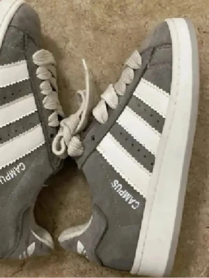Grå Campus sneakers med vita ränder - Snygga grå Adidas Campus sneakers med klassiska vita ränder på sidorna och vit sula. Ovandelen är i mocka och skorna har vita skosnören. Perfekta för en streetwear-look och passar till det mesta i garderoben.