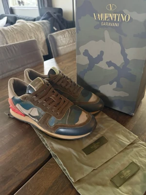 Valentino Rock Runners  - Rock Runners från Valentino Garavani med camomönster i grönt, blått och beige. Skorna har bruna mockadetaljer, röda nitar vid hälen och vita samt svarta inslag. Klassisk låg modell med bruna skosnören, perfekt för både vardag och finare tillfällen. Jättebra skick och säljer då jag har växt ur dom. Storleken är 41 men passar även 42. Hör av er vid funderingar. Kom gärna med bud😁