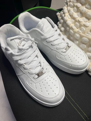 Nike Air Force 1 vita sneakers - Klassiska Nike Air Force 1 sneakers i helvitt läder med perforerad tå, vita skosnören och silverfärgad detalj vid snörningen. Snygg och tidlös modell med låg profil och diskret Nike-logga på sidan och hälen. Perfekta för en clean och stilren look.