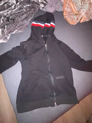 Svart Moncler hoodie med dragkedja - Svart hoodie från Moncler med dragkedja och huva. Huven har röda, vita och blå ränder som ger en snygg kontrast. Moncler-logga på ärmen. Perfekt för dig som vill ha en stilren och exklusiv look. Materialet är mjukt och bekvämt.