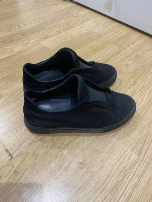 Svarta sneakers i mocka-look - Stilrena svarta sneakers med låg profil och ovandel i mocka-liknande material. Skorna har en enkel design utan synliga snören och en diskret sula i grått. Perfekta för dig som gillar minimalistisk stil.