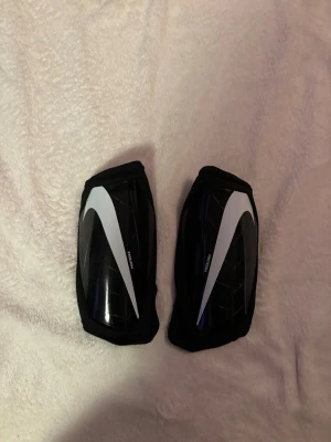 Nike benskydd svart/vit - Säljer ett par svarta benskydd från Nike med vit swoosh-logga. Skydden har en hård yta framtill och meshmaterial på baksidan för ventilation. Perfekta för fotboll eller andra sporter där du behöver extra skydd för benen.