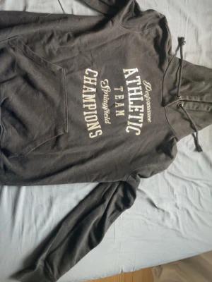 Svart hoodie från PXS Active Sports - Svart hoodie med tryck i vitt framtill där det står 'Performance Athletic Team Champions Springfield'. Har huva med dragsko och en stor magficka. Perfekt för en sportig och avslappnad stil.