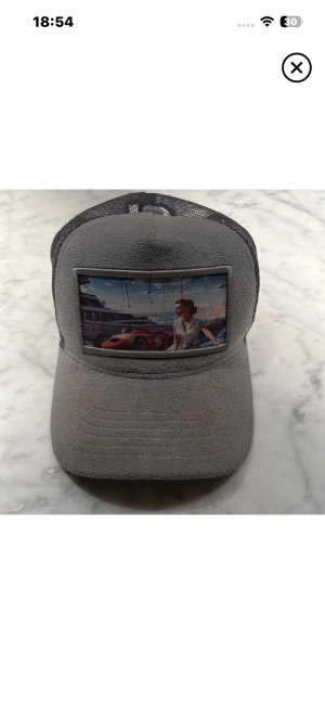 Grå meshkeps med bilmotiv - Säljer en grå keps med mesh baktill och justerbar snapback. Framtill finns ett färgglatt tryck med en sportbil och en person framför en byggnad. Kepsen har böjd skärm och är tillverkad i tyg och mesh för skön ventilation.