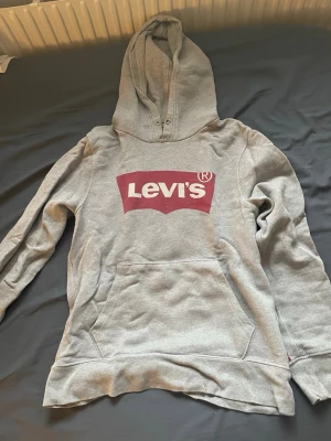 Grå Levi's hoodie med logga XS - Säljer en grå hoodie från Levi's i storlek XS. Tröjan har stor röd Levi's-logga på bröstet, klassisk känguruficka och huva med dragsko. Tillverkad i mjukt sweatshirtmaterial, perfekt för en avslappnad stil.