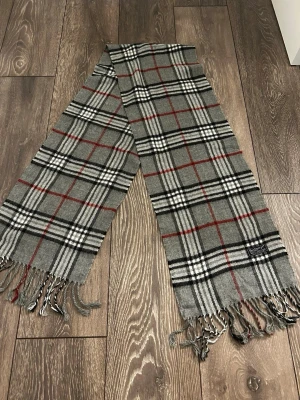 Grå rutig halsduk från Burberry - Klassisk halsduk i grått med svart, vitt och rött rutmönster från Burberry. Halsduken har fransar i ändarna och är tillverkad i mjukt material som känns lyxigt. Perfekt accessoar för att lyfta din outfit med en tidlös design.