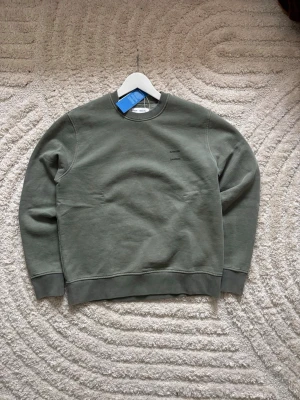 Med prislapp/Grön sweatshirt från Samsøe Samsøe  - Stilren grön sweatshirt från Samsøe Samsøe med diskret logga på bröstet. Tröjan har rund halsringning, ribbade muddar vid ärmslut och nederkant samt en avslappnad passform. Perfekt till jeans eller cargo.