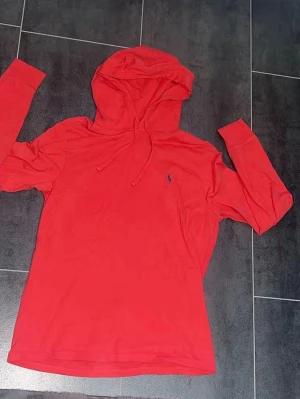 Röd hoodie från Polo Ralph Lauren - Snygg röd hoodie från Polo Ralph Lauren med klassisk huva och dragsko. Liten blå logga broderad på bröstet. Tillverkad i mjukt och lätt material, perfekt för en avslappnad stil. Långärmad modell som passar året runt.