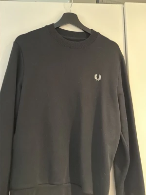 Svart sweatshirt från Fred Perry - Klassisk svart sweatshirt från Fred Perry med rund halsringning och vit broderad logga på bröstet. Tröjan har ribbade muddar vid ärmslut och nederkant. Perfekt för en stilren och enkel look.