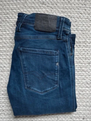 Mörkblå jeans från Replay Anbass - Snygga mörkblå jeans från Replay med klassisk femficksdesign och läderpatch bak i midjan. Jeansen har smal passform och är tillverkade i stretchigt denimtyg för extra komfort. Perfekta för en trendig och avslappnad look. Modellen är Anbass. Storlek 30/32 