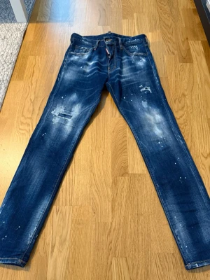 Dsquared2 blå distressed jeans - Säljer ett par blå jeans från Dsquared2 med slim fit och coola distressed detaljer som slitningar och färgstänk. Jeansen har klassisk femficksmodell och är tillverkade i bomull. Perfekta för en trendig och avslappnad stil.