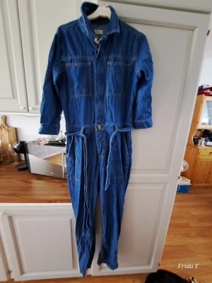 Blå denim jumpsuit med knytband - Snygg blå denim jumpsuit med långa ärmar, dragkedja framtill och knytband i midjan. Två bröstfickor och två sidofickor ger en cool och avslappnad look. Perfekt för dig som gillar streetstyle och vill ha något bekvämt men trendigt.