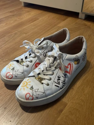 Vita sneakers med print från Philip Hog - Limited edition vita sneakers från Philip Hog med färgglada prints som sol, hjärta, fredsduva och texten 'I love Stockholm'. Skorna har vita snören, guldfärgade detaljer och är tillverkade i läder. Perfekta för dig som vill sticka ut med din stil.