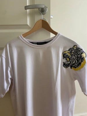 Vit t-shirt med tigertryck från Philipp Plein - Snygg vit t-shirt från Philipp Plein med ett stort tigertryck i svart och gult på axeln. T-shirten har rund hals och korta ärmar. Tillverkad i mjuk bomull för en bekväm känsla. Perfekt för dig som vill sticka ut med en unik detalj.