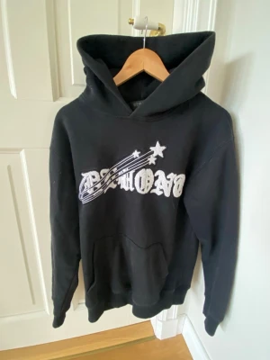 Svart hoodie från Demont med tryck - Svart hoodie från Demont med stor huva och ett fett vitt tryck med stjärna och texten 'DEMONT' på bröstet. Klassisk känguruficka framtill och ribbade muddar. Perfekt för dig som gillar streetwear och vill sticka ut lite extra.