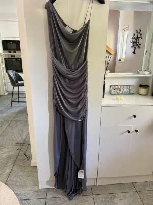 Glittrig silver långklänning - Snygg långklänning i glittrigt silver med draperad framsida och tunna axelband. Klänningen har en elegant omlottkänsla och ett mjukt, följsamt material som ger ett lyxigt intryck. Perfekt för dig som vill sticka ut på festen.