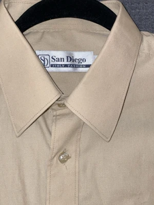 Beige skjorta från San Diego - Klassisk beige skjorta från San Diego med korta ärmar och bröstficka. Skjortan har en stilren design med knappar framtill och en mjuk krage. Perfekt för dig som gillar enkel och clean stil.